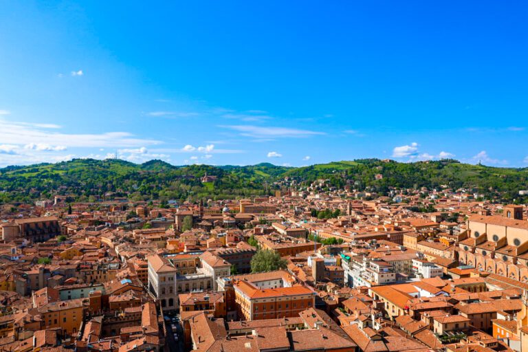 Panoramic view of Bologna, Italy, showcasing the historic city center with its iconic red-tiled rooftops and medieval architecture against a clear blue sky
